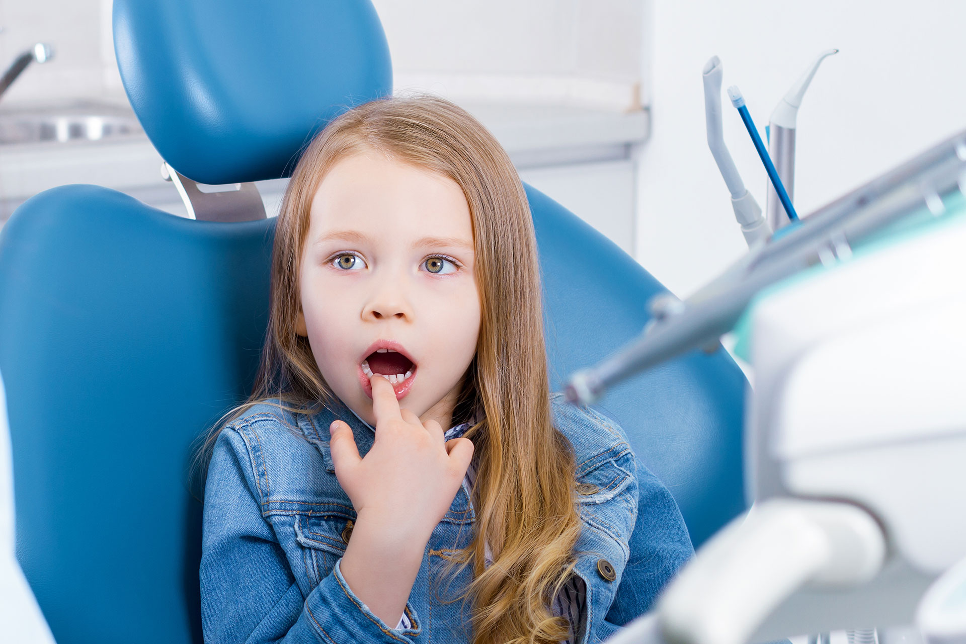 Young girl with open mouth, seated in dental chair, surrounded by dental equipment.