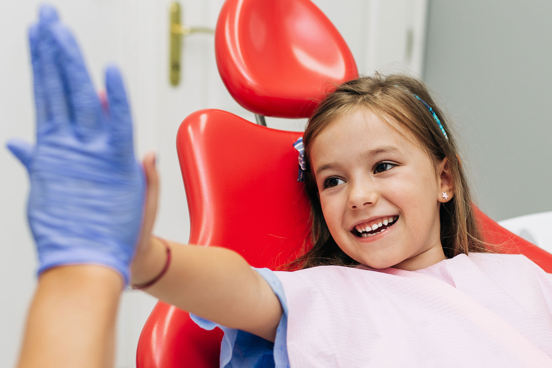 A young girl is giving a thumbs-up while wearing gloves and sitting in a dental chair.