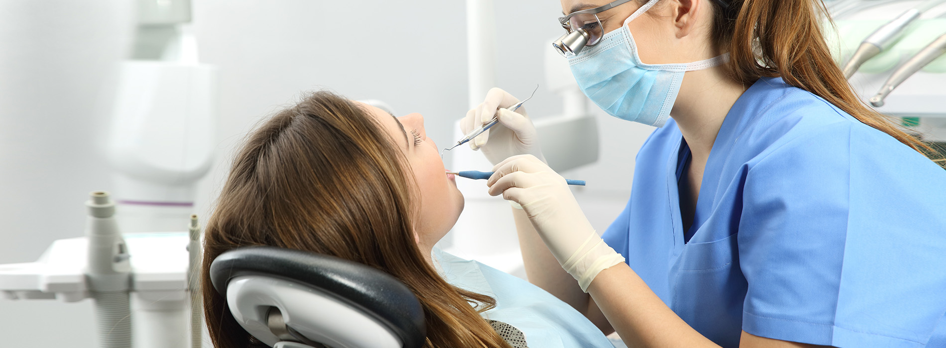 A dental hygienist is attending to a patient in a dental office, with the patient seated in an examination chair and receiving care.
