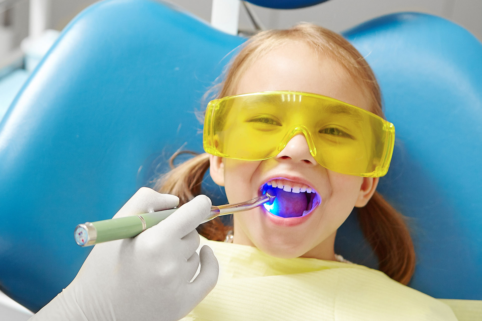 A young girl in a dental office, wearing yellow protective goggles and holding a toothbrush, brushing her teeth while seated in a dentist s chair.