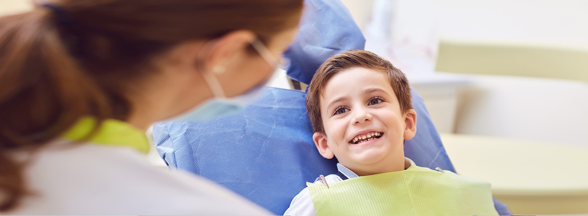 A young boy sitting in a dentist s chair, smiling and looking at the camera, while a dental professional attends to him.