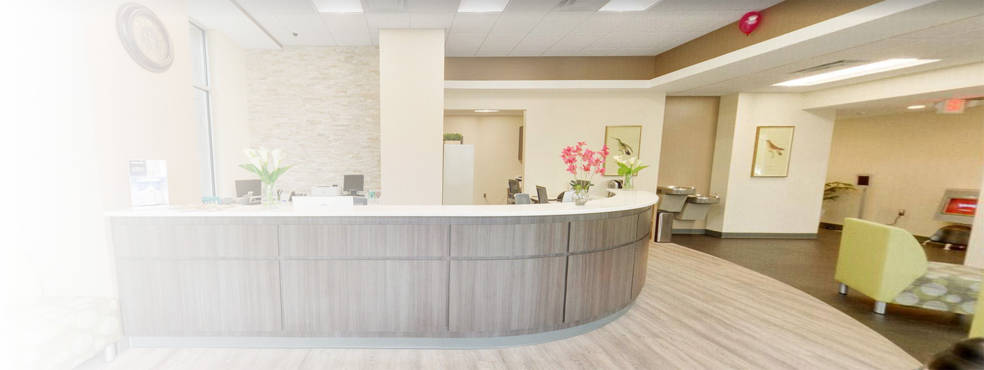 The image shows an interior view of a modern lobby or waiting area within a building, featuring a reception desk with a curved countertop, a seating area with a large window, and a contemporary design with neutral colors.
