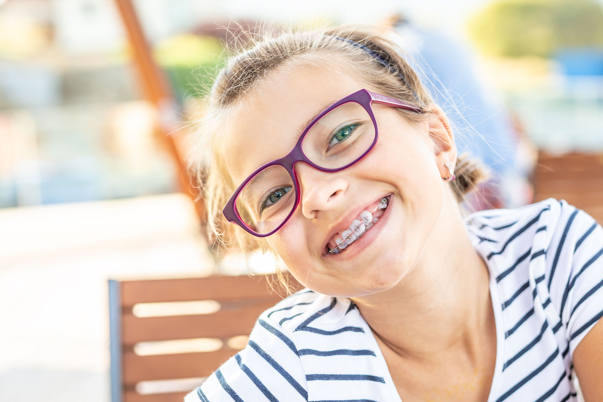 Girl in purple glasses smiling at camera, seated outdoors.