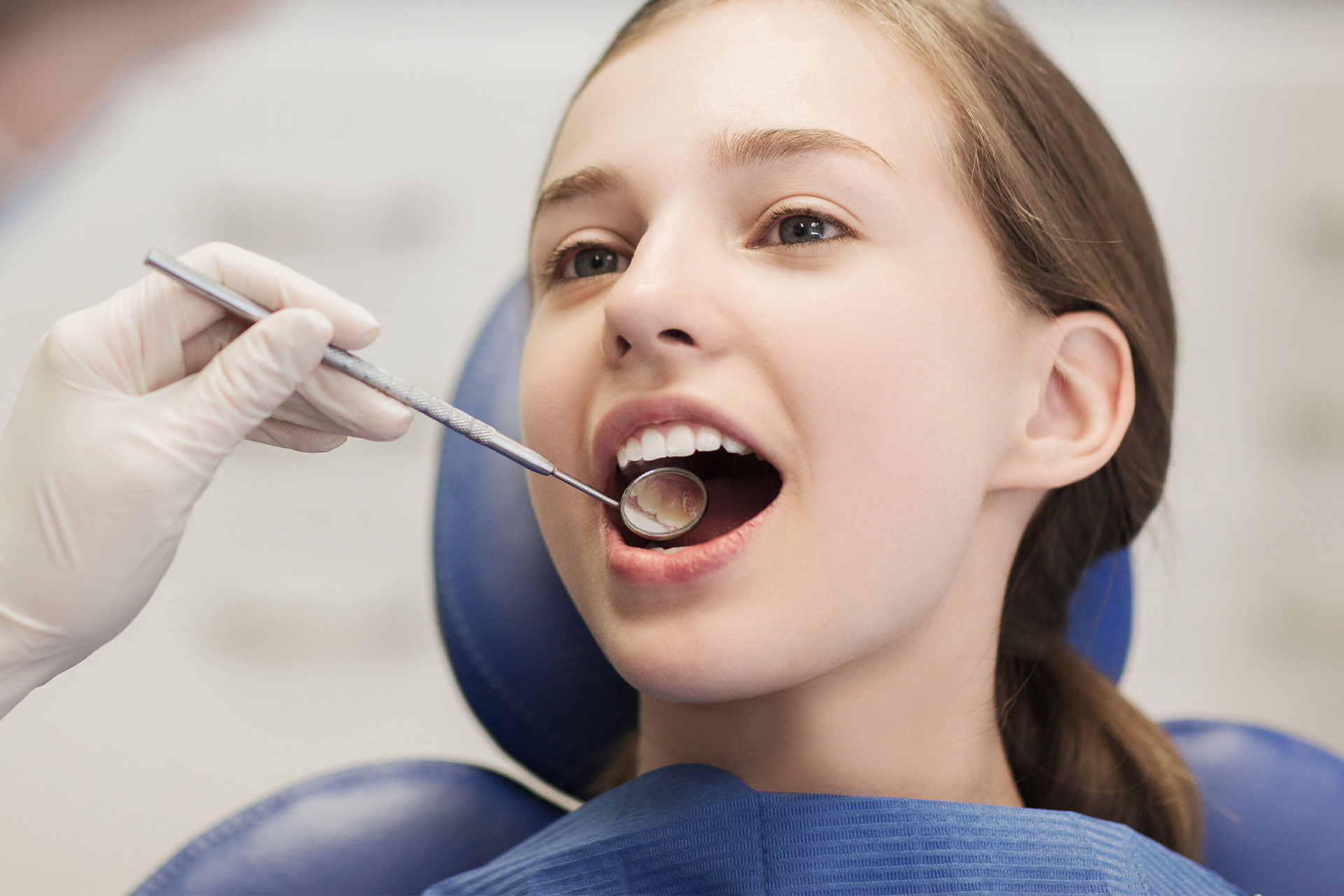 The image shows a young woman in a dental office setting, receiving dental care. She is seated in a dental chair with her mouth open, wearing a blue surgical mask and holding a small object, possibly a toothbrush or dental tool. A dentist is seen in the background, wearing gloves and working at a counter with various dental instruments.