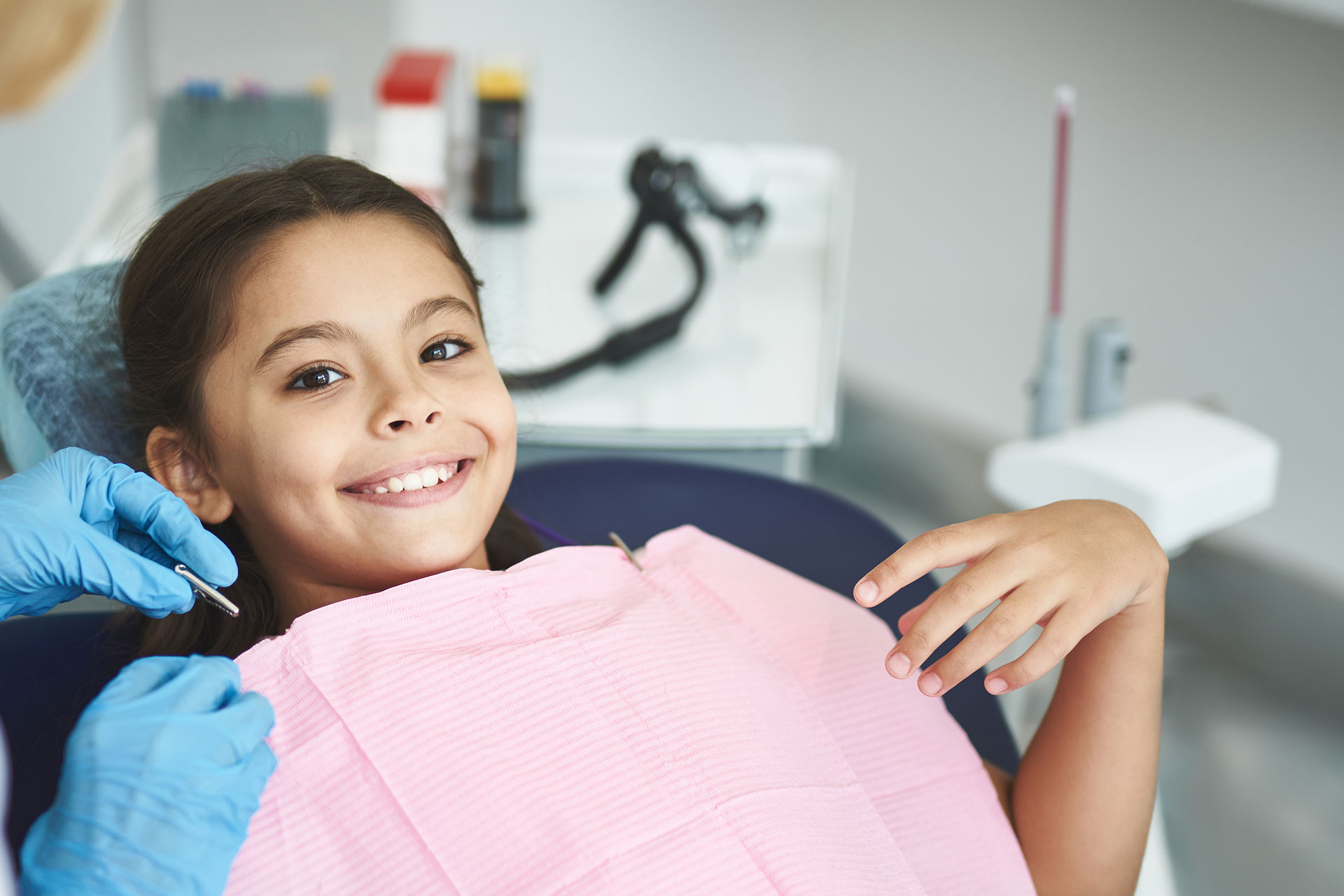 The image shows a young girl sitting in a dental chair, smiling at the camera, with a dentist standing behind her wearing gloves and holding a mirror.