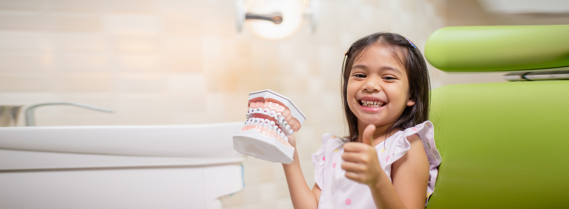 A young girl is standing in front of a dental chair, holding a toothbrush and smiling.