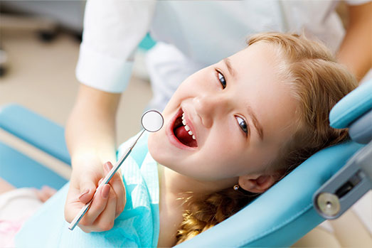 A young girl is sitting in a dental chair, smiling broadly while holding a toothbrush and looking at the camera.