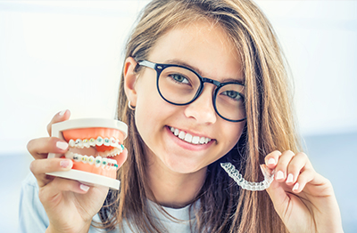 The image shows a young woman smiling at the camera, holding a toothbrush with bristles and a toothpaste tube in her other hand. She is wearing glasses and has a small white gap between her front teeth.
