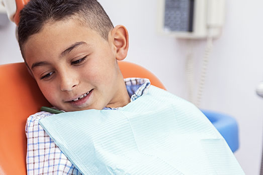 A young boy in a dental chair, wearing a blue shirt and smiling, with his teeth being checked or cleaned by a dental professional.
