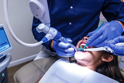 The image shows a dental professional working on a patient s teeth using a dental drill, with the patient seated in a dental chair and wearing protective eyewear.