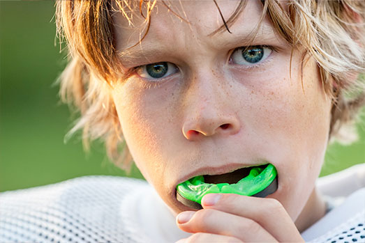 The image shows a young boy with blonde hair, wearing a football uniform and holding a green toothbrush in his mouth.