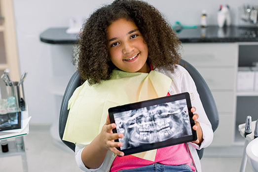 A young girl holding a tablet in front of her face, smiling at the camera, while sitting in a dental chair.