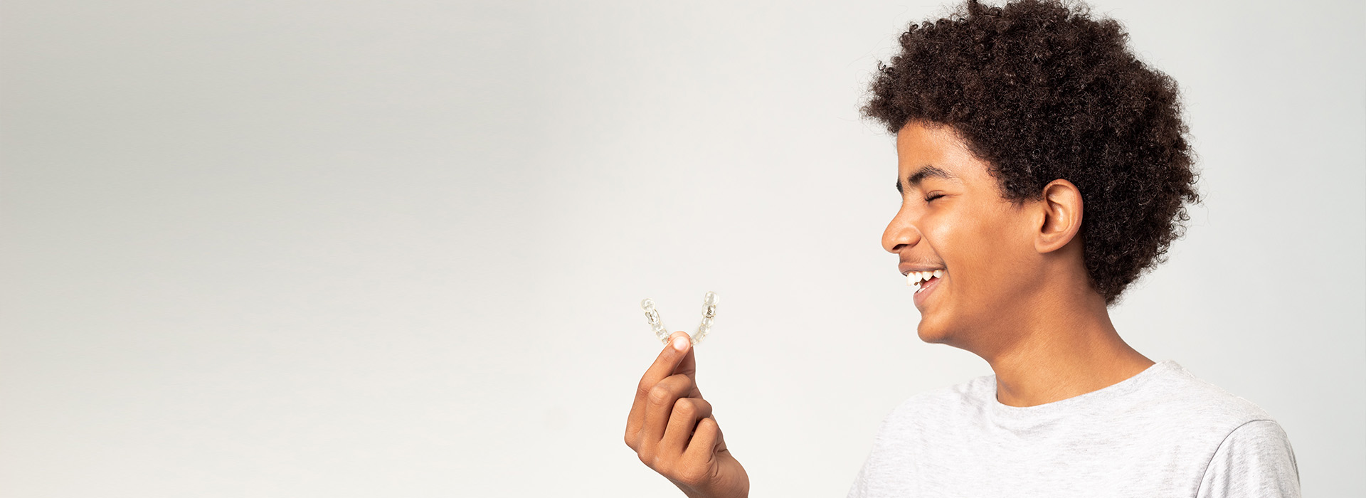 A young person with short hair, wearing a white shirt, is smiling and holding a small object that resembles a flower or star in their hand. The background is plain and light-colored, emphasizing the subject in the foreground.