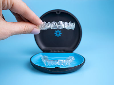 A person s hand holds a clear, plastic dental retainer with braces inside an open blue case.