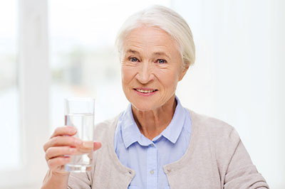 The image shows a woman, presumably older due to her appearance and attire, holding a glass of water. She is smiling and appears to be in a good mood or making a positive gesture.