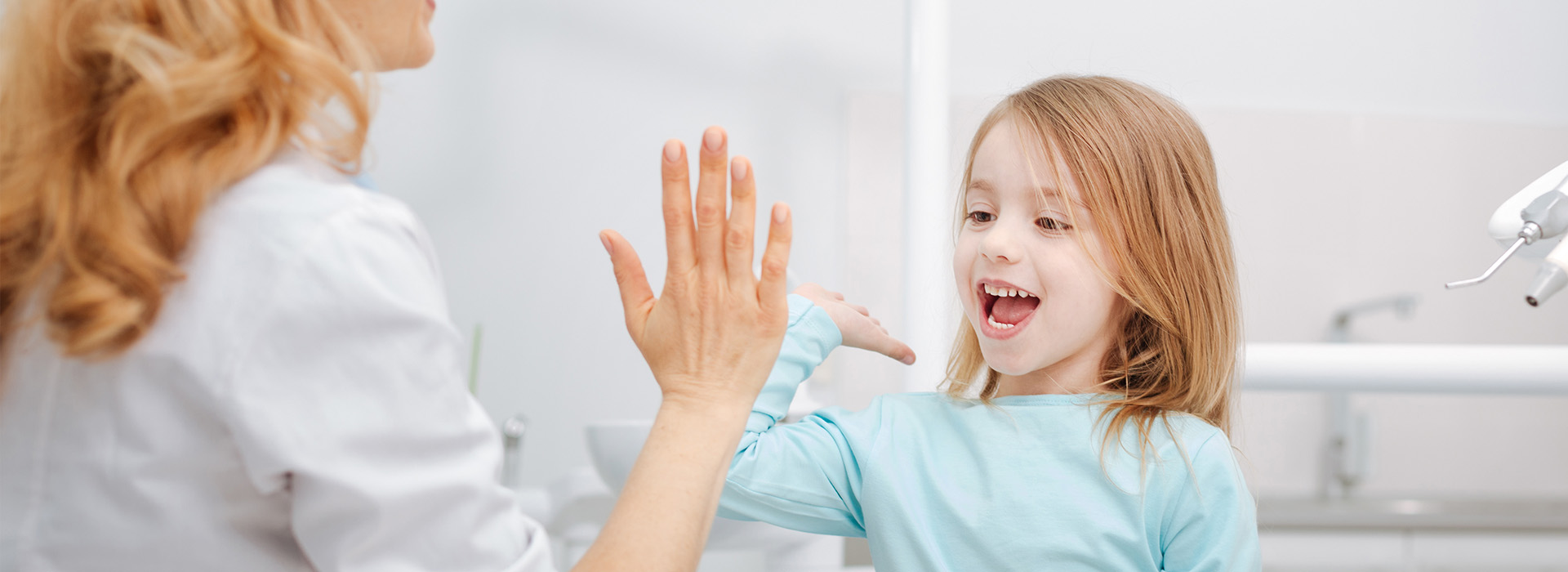 A woman and a young girl are seen in the image, with the woman holding up her hand towards the child. The girl appears to be smiling or laughing, while the woman is looking at her with a smile on her face. They seem to be in an indoor setting, possibly a bathroom, as suggested by the presence of a sink and mirror in the background.