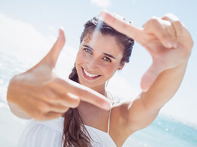 The image shows a person taking a selfie with one hand, smiling and looking directly at the camera. They are outdoors in bright sunlight, wearing a white top, and there is an oceanic backdrop.