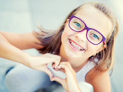 A young girl with glasses, making a heart shape with her hands against a blurred background.